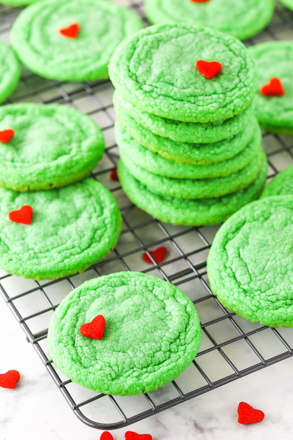 Green Grinch Cookies topped with a small heart shaped sprinkle spread out over a cooling rack on a white table top