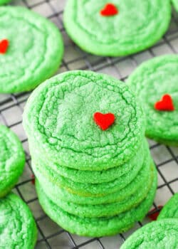 Green Grinch Cookies topped with a small heart shaped sprinkle spread out over a cooling rack on a white table top