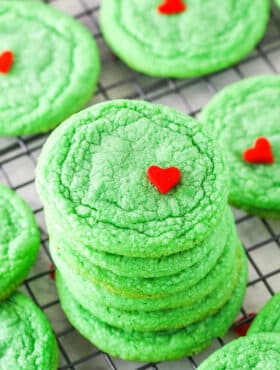 Green Grinch Cookies topped with a small heart shaped sprinkle spread out over a cooling rack on a white table top