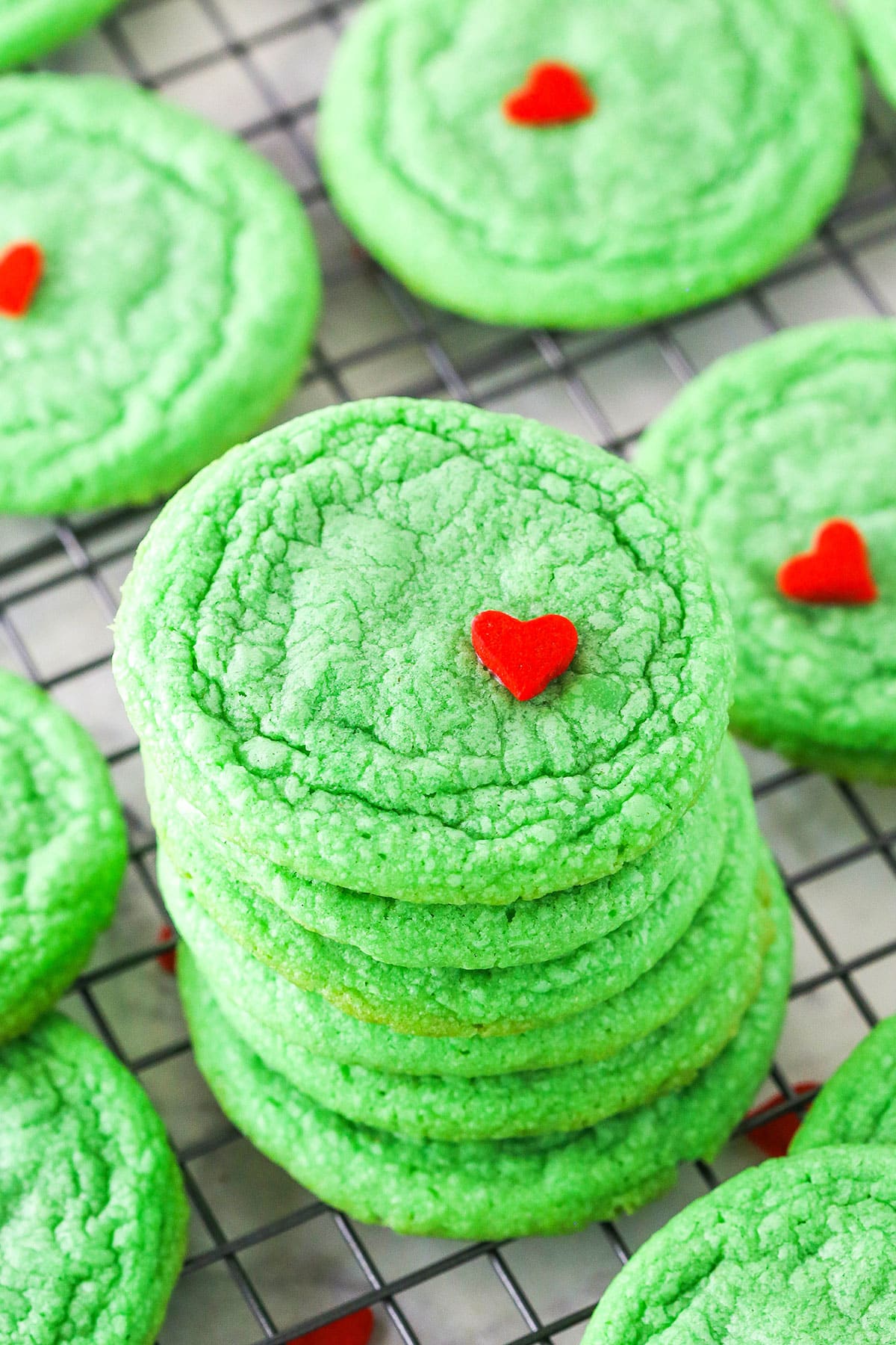 Green Grinch Cookies topped with a small heart shaped sprinkle spread out over a cooling rack on a white table top