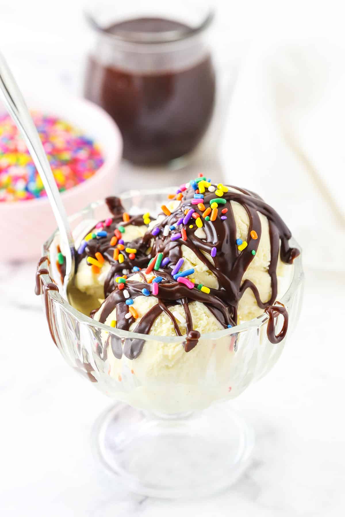 Homemade Hot Fudge Sauce drizzled over three scoops of vanilla ice cream and topped with multicolored sprinkles in a glass bowl with a spoon