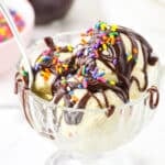 Homemade Hot Fudge Sauce drizzled over three scoops of vanilla ice cream and topped with multicolored sprinkles in a glass bowl with a spoon