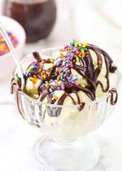 Homemade Hot Fudge Sauce drizzled over three scoops of vanilla ice cream and topped with multicolored sprinkles in a glass bowl with a spoon