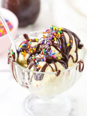 Homemade Hot Fudge Sauce drizzled over three scoops of vanilla ice cream and topped with multicolored sprinkles in a glass bowl with a spoon
