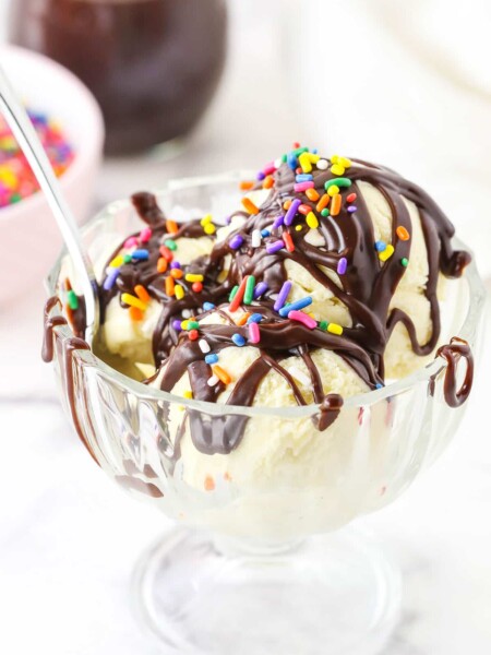 Homemade Hot Fudge Sauce drizzled over three scoops of vanilla ice cream and topped with multicolored sprinkles in a glass bowl with a spoon