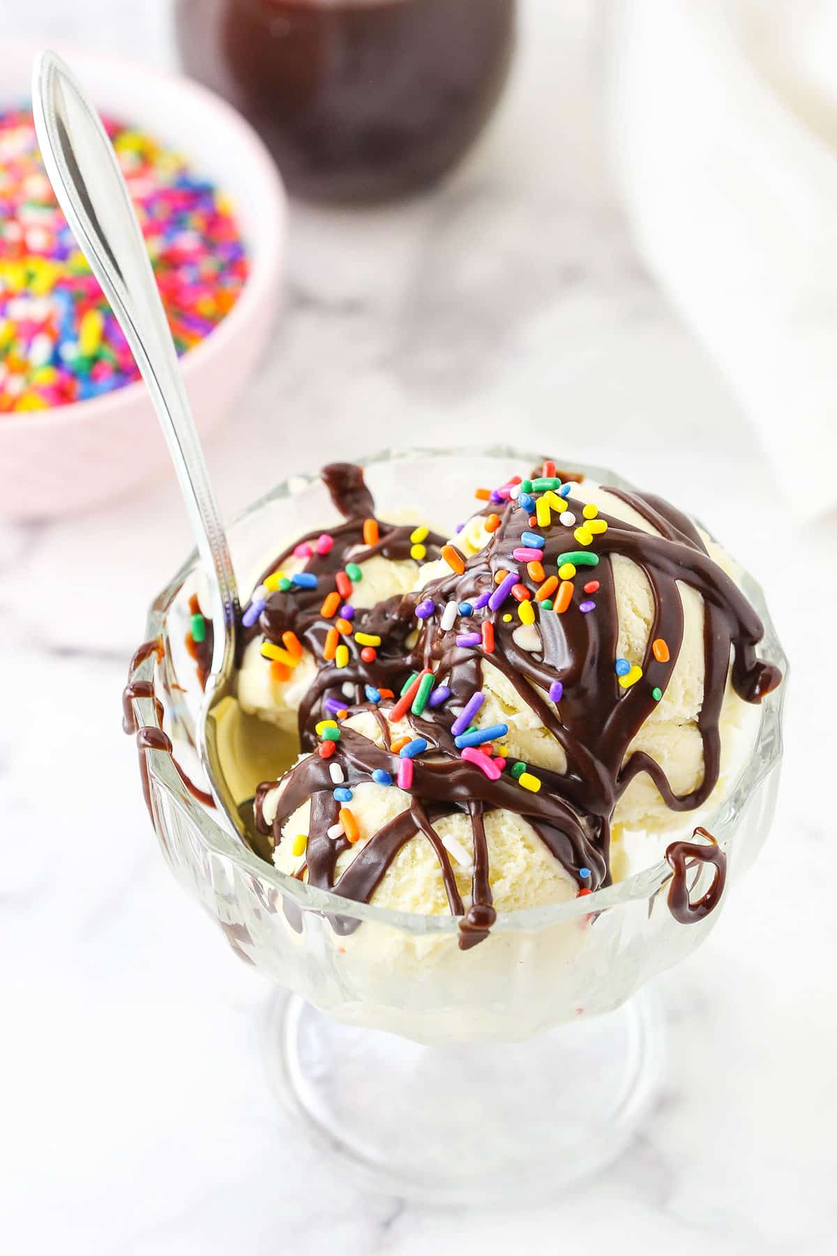 Homemade Hot Fudge Sauce drizzled over three scoops of vanilla ice cream and topped with multicolored sprinkles in a glass bowl with a spoon