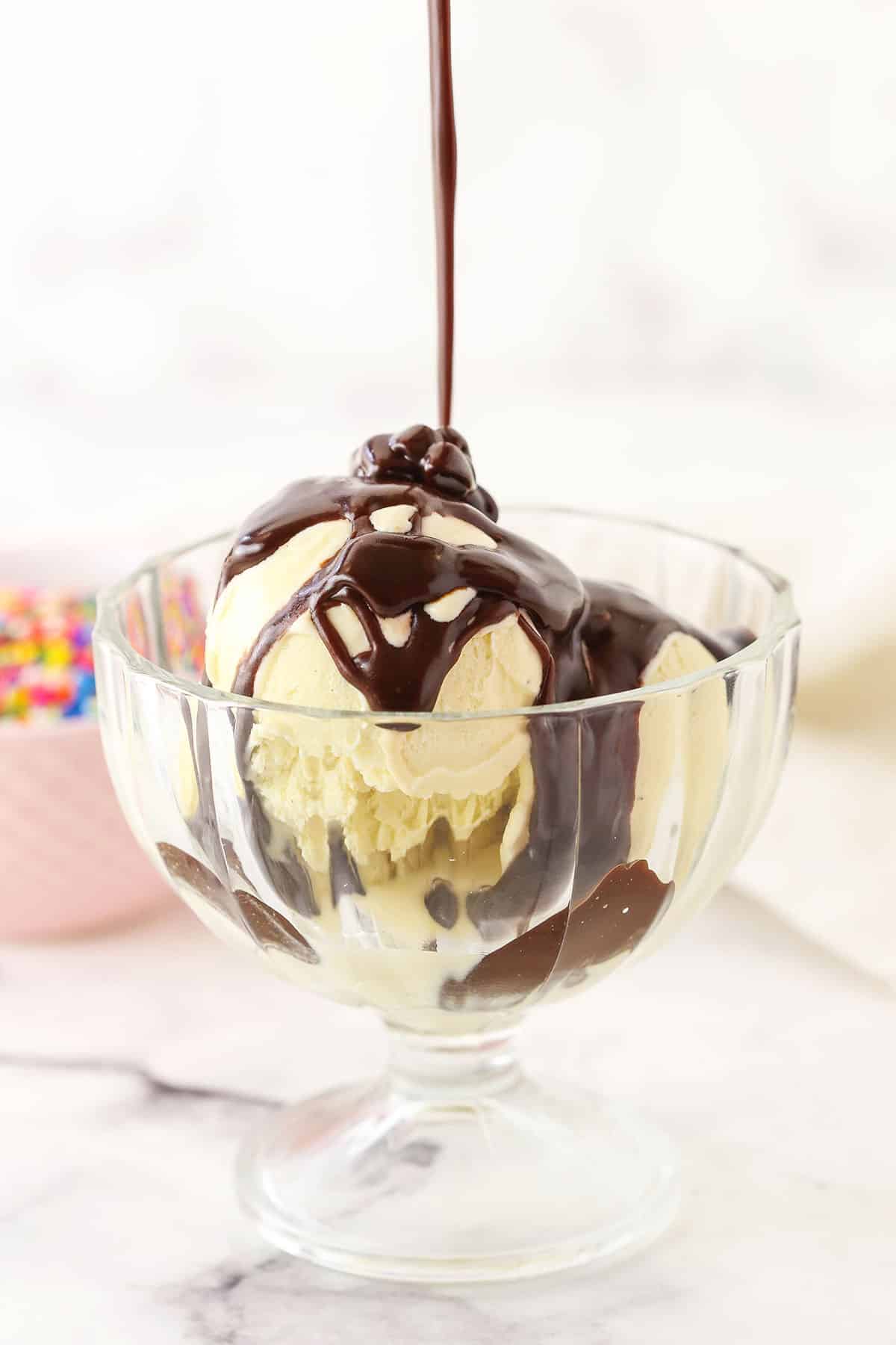 Homemade Hot Fudge Sauce being drizzled over three scoops of vanilla ice cream in a glass bowl with a spoon
