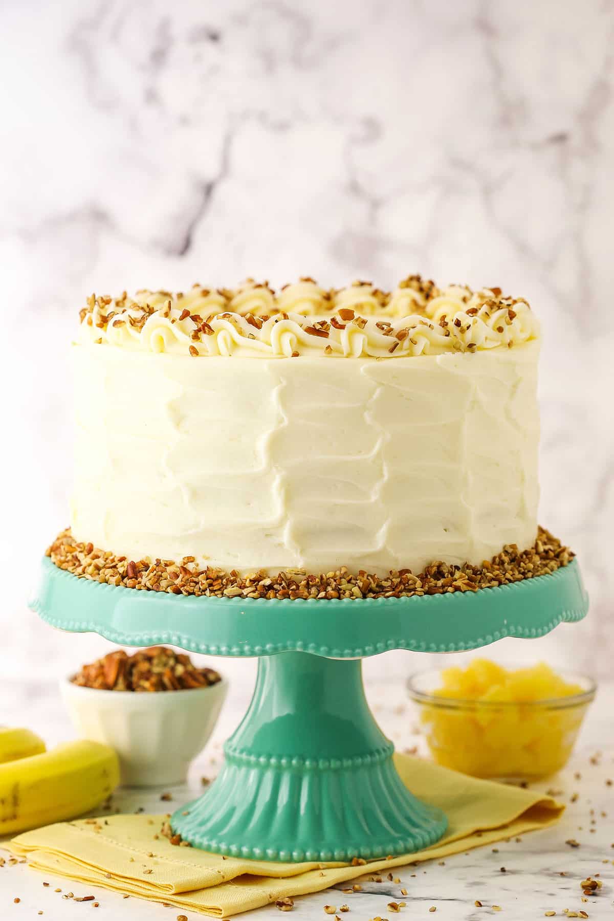Side view of a full Hummingbird Cake topped with white swirls and chopped pecans on a blue cake stand