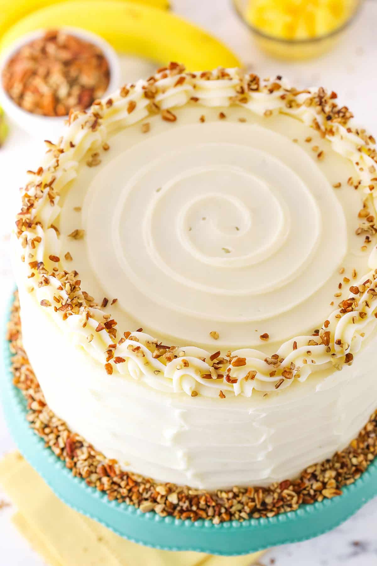 Overhead view of a full Hummingbird Cake topped with white swirls and chopped pecans on a blue cake stand