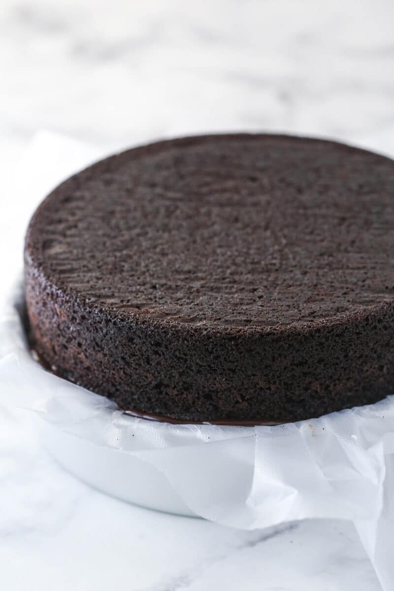 A chocolate cake layer on top of a fudge and cookie crunchies layer on top of an ice cream layer in a cake pan lined with clear wrap.