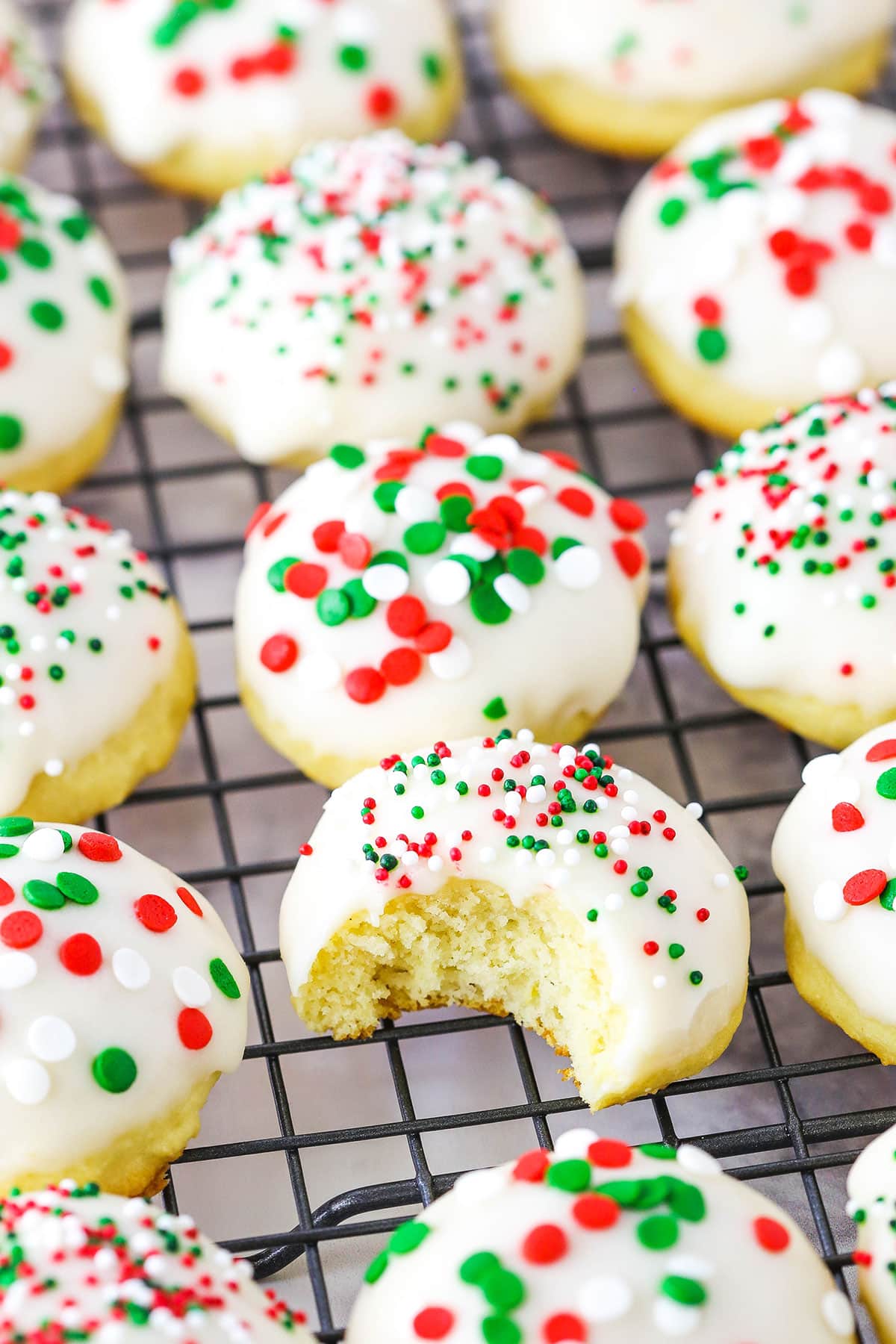 Italian ricotta cookies are soft, cake-like cookies with subtle tangy lemon flavor and sweet icing. They are perfect for any holiday! Italian ricotta cookies with red, green, and white sprinkles spread over a metal cooling rack with one cookie missing a bite.