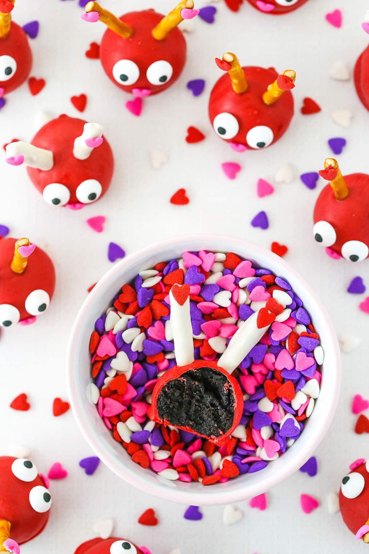Love Bug Oreo Cookie Ball with a bite taken out in a white bowl full of heart shaped sprinkles with more Love Bug Oreo Cookie Balls in the background