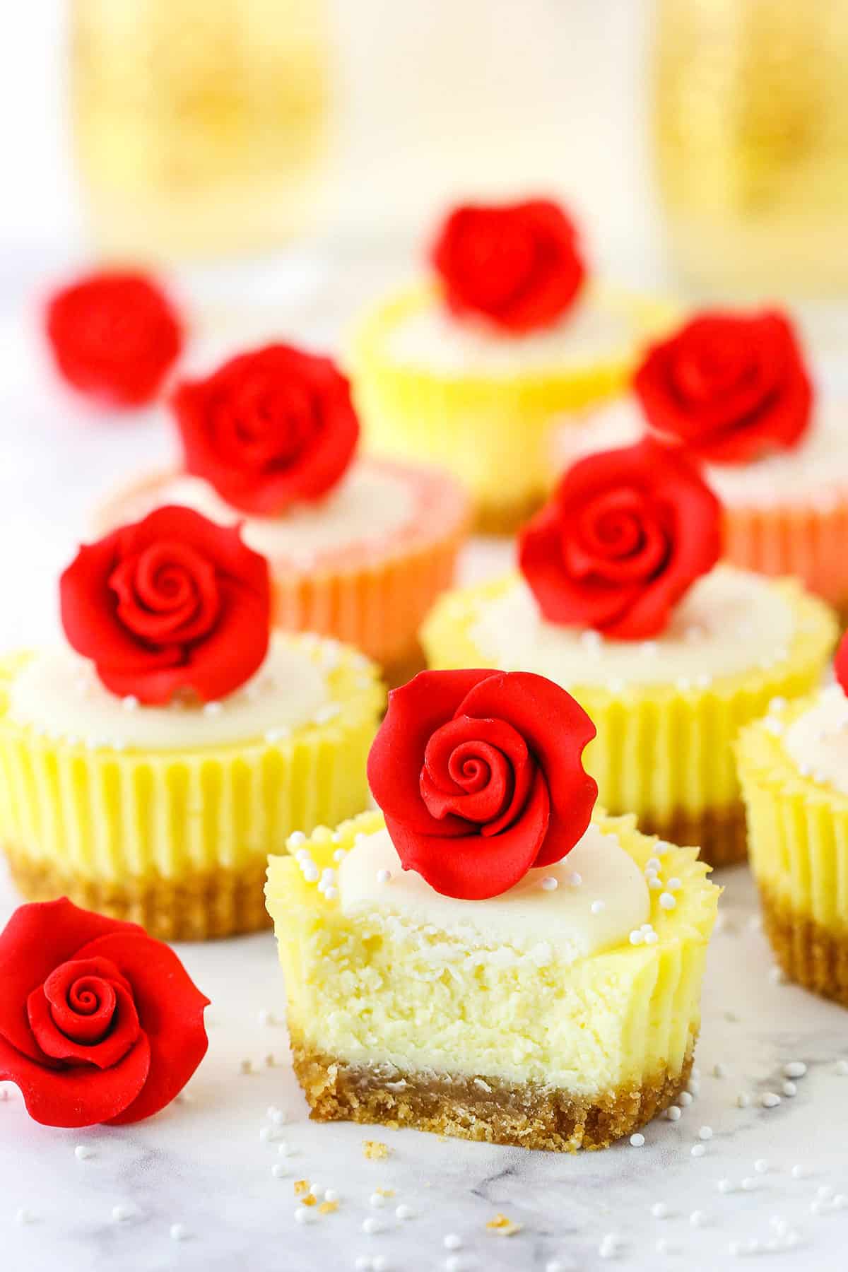 Mini Rose Cheesecakes topped with white icing and a red rose with the front Mini Rose Cheesecake missing a bite on a white marble table