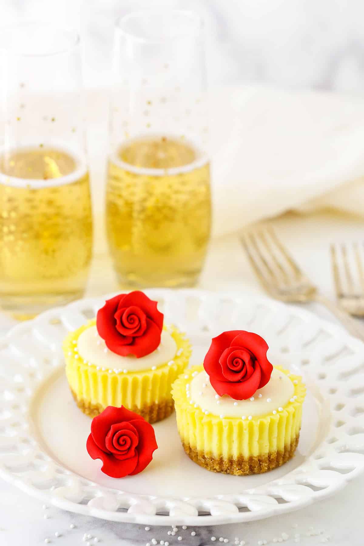 Two Mini Rose Cheesecakes topped with white icing and a red rose on a white plate next to another rose with two glasses of champagne in the background