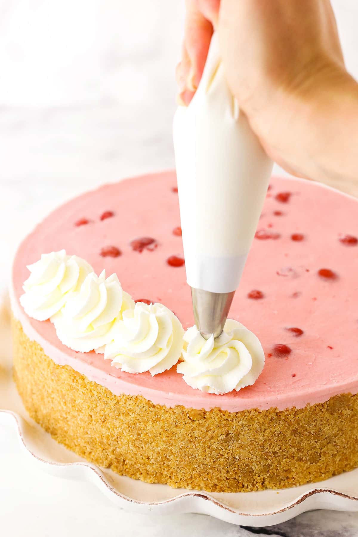 A step in making No Bake Cherry Almond Cheesecake showing decorating the cheesecake with the almond whipped cream swirls using a piping bag and tip.