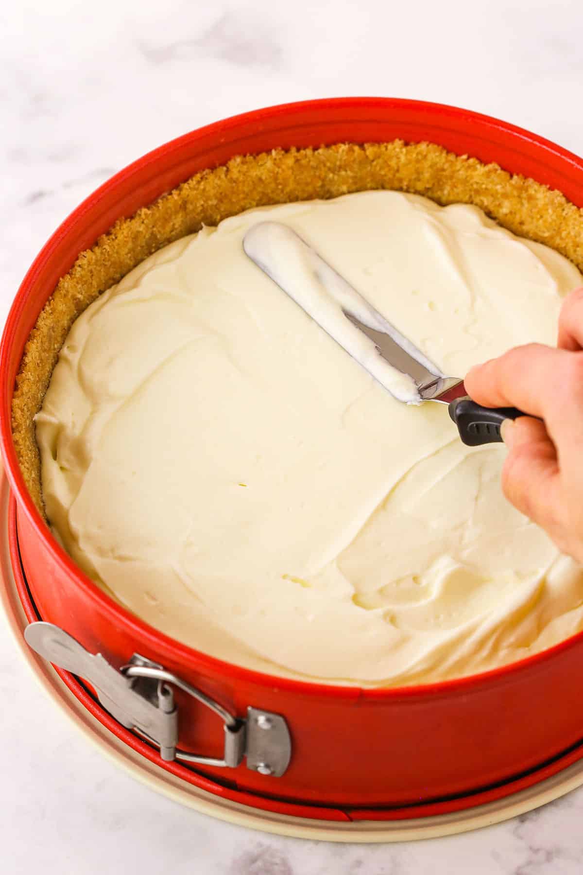 A step in making No Bake Cherry Almond Cheesecake showing spreading the almond filling evenly into the chilled crust, in the springform pan, using an offset spatula