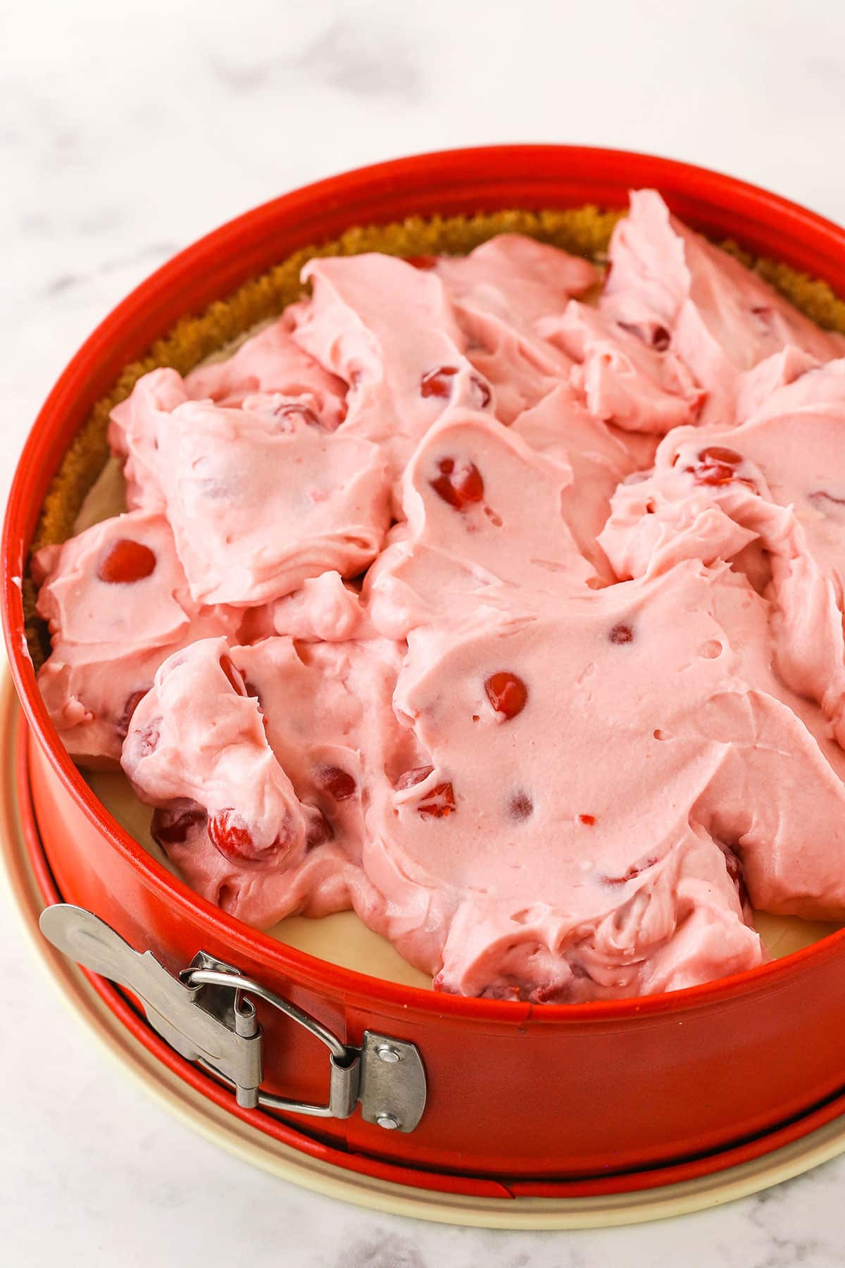 A step in making No Bake Cherry Almond Cheesecake showing adding the cherry mixture over top of the almond filling in the springform pan.