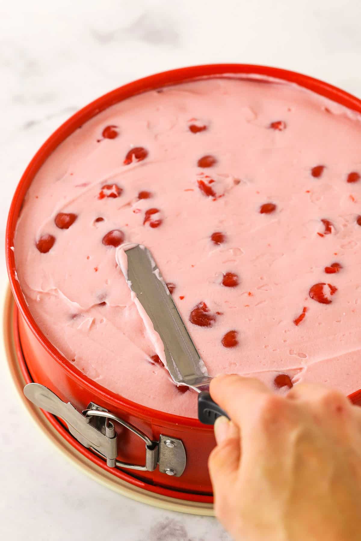 A step in making No Bake Cherry Almond Cheesecake showing spreading the cherry mixture into an even layer using an offset spatula