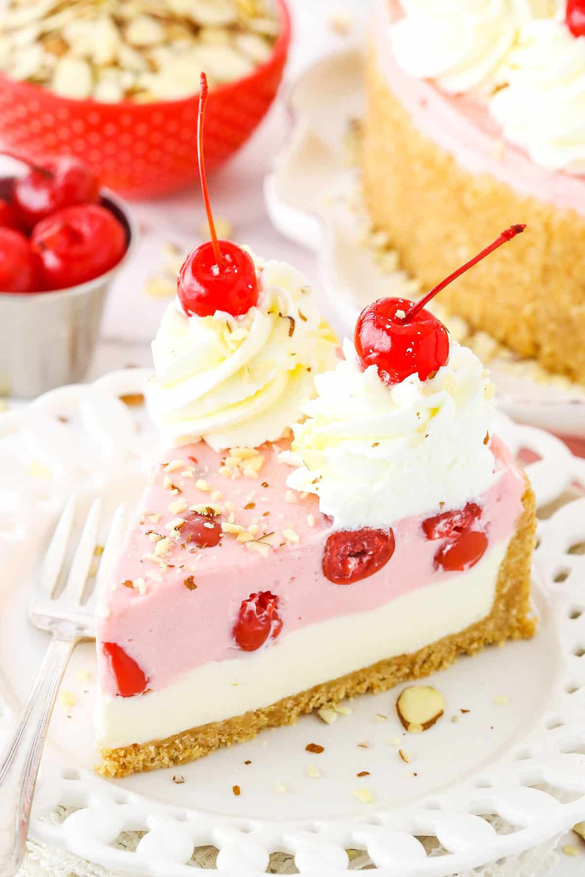 This No Bake Cherry Almond Cheesecake is super simple to make and FULL of flavor! It's got a vanilla cookie crust and layers of delicious almond and cherry cheesecake! A slice of No Bake Cherry Almond Cheesecake topped with white swirls and cherries on a white plate with a fork