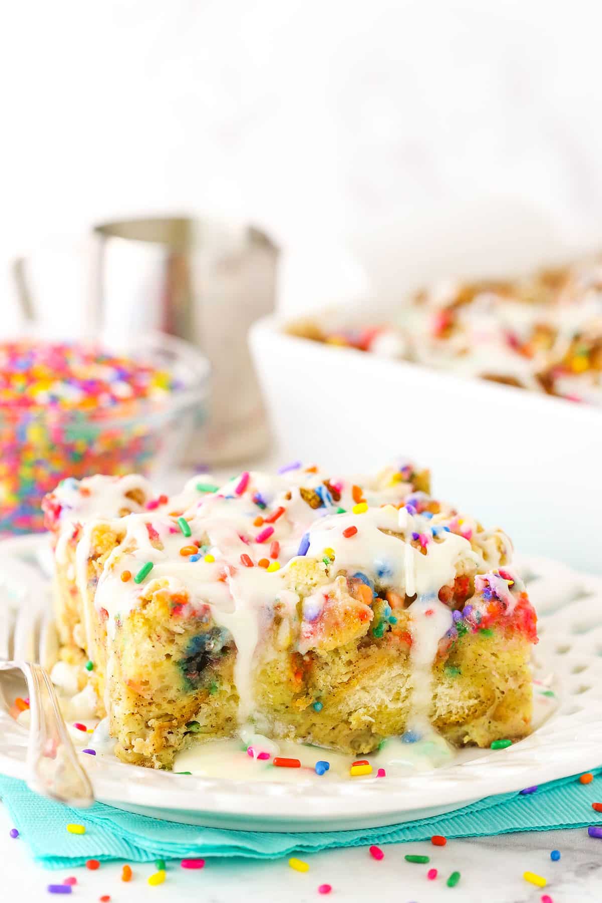 Side view of a slice of Overnight Birthday Cake French Toast Casserole topped with colorful sprinkles and icing on a white plate with a fork