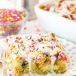 Side view of a slice of Overnight Birthday Cake French Toast Casserole topped with colorful sprinkles and icing on a white plate with a fork