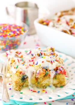 Side view of a slice of Overnight Birthday Cake French Toast Casserole topped with colorful sprinkles and icing on a white plate with a fork