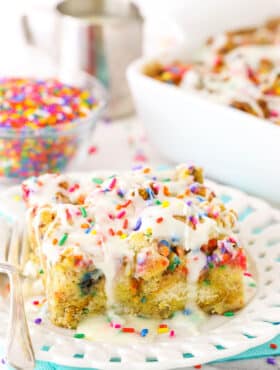 Side view of a slice of Overnight Birthday Cake French Toast Casserole topped with colorful sprinkles and icing on a white plate with a fork