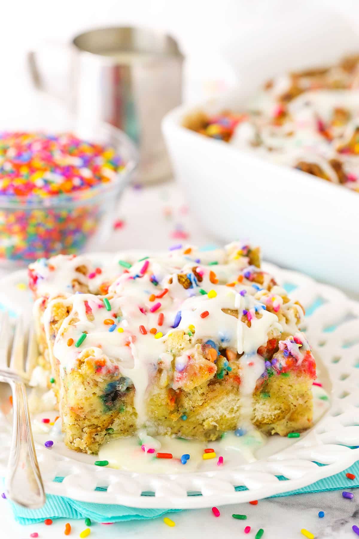 Side view of a slice of Overnight Birthday Cake French Toast Casserole topped with colorful sprinkles and icing on a white plate with a fork