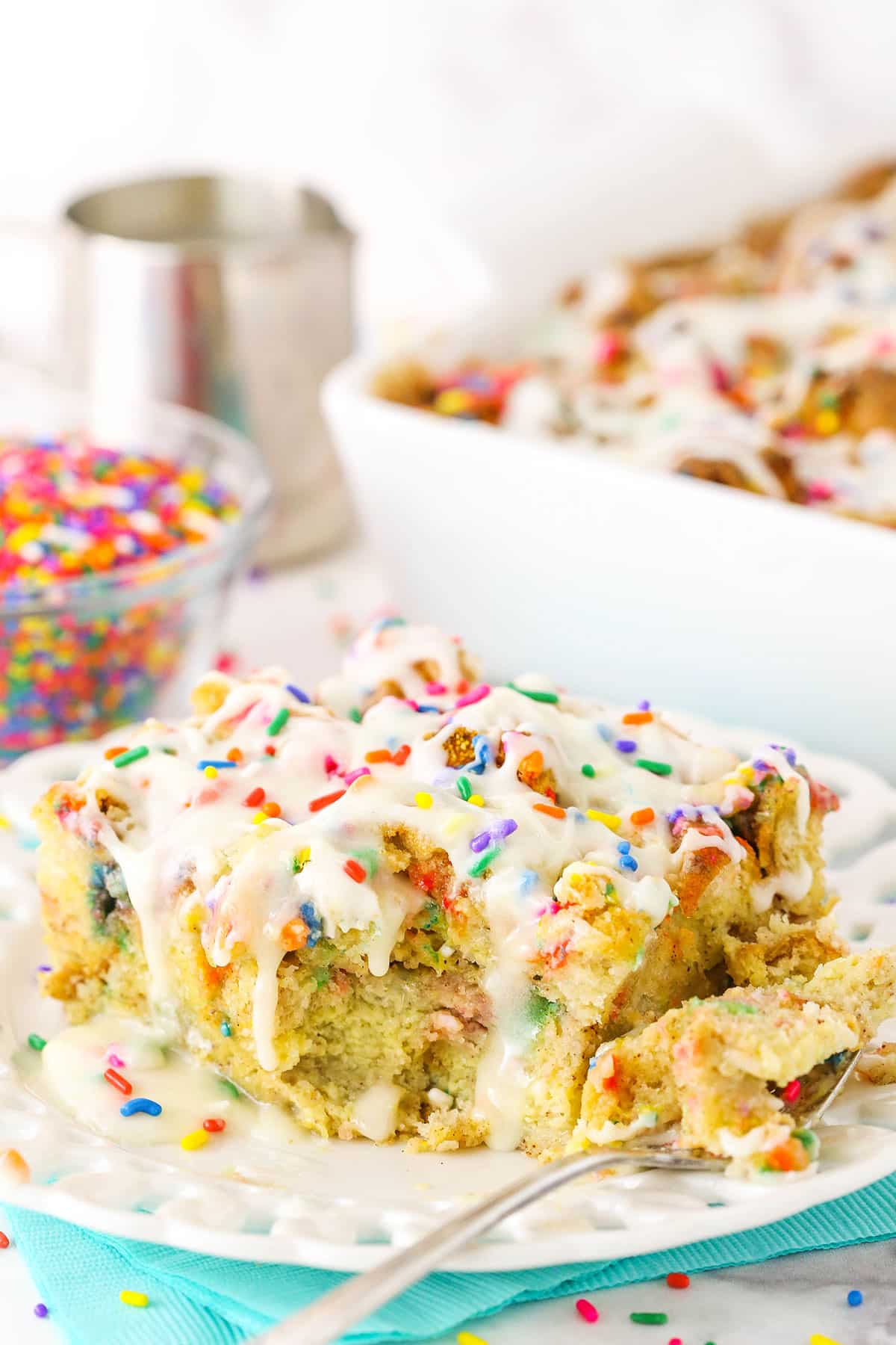 Side view of a slice of Overnight Birthday Cake French Toast Casserole topped with colorful sprinkles and icing with a bite removed on a white plate with a fork