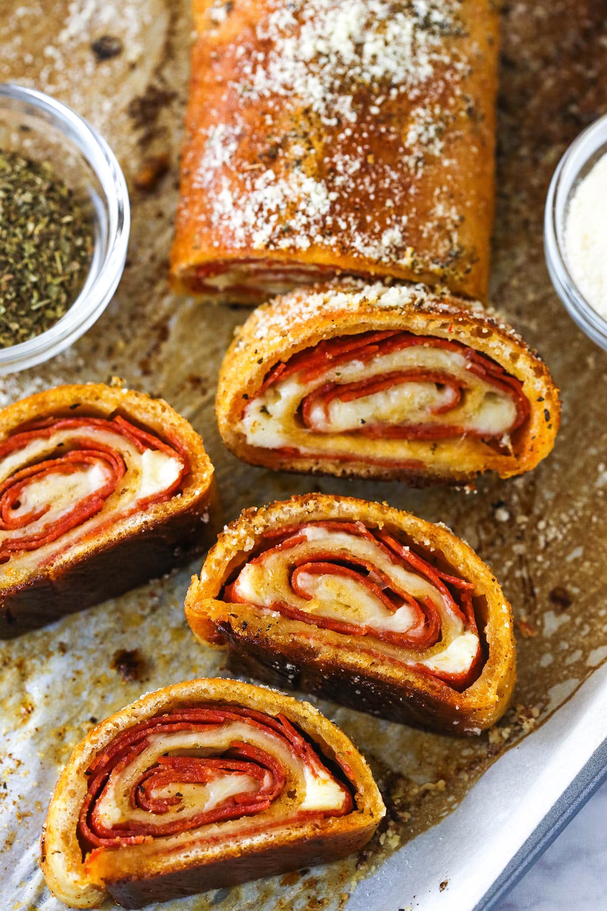 A full pepperoni roll with slices of pepperoni roll near a small bowl of Italian seasoning and a small bowl of cheese.