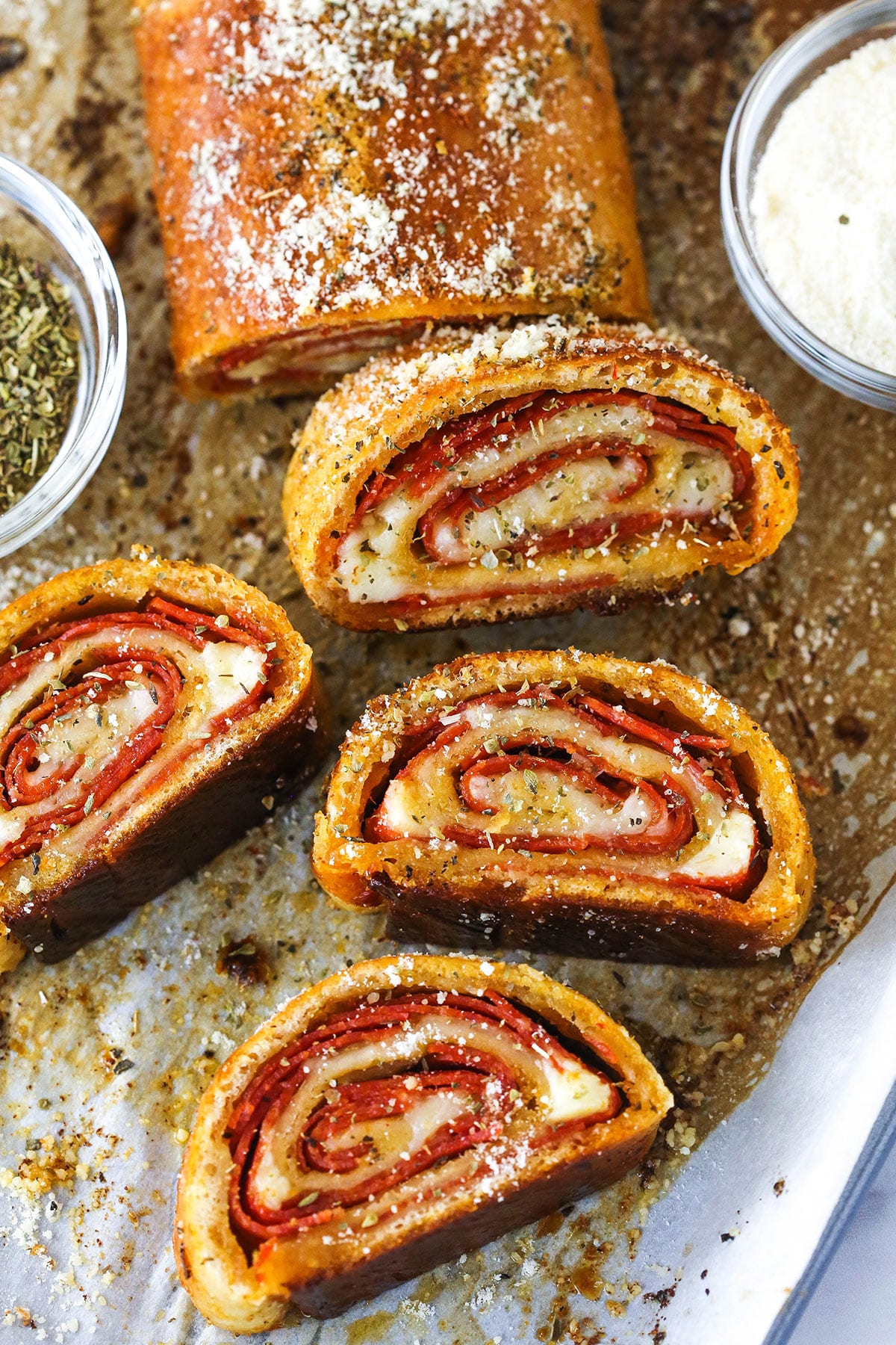 Pepperoni rolls are made up of pizza dough brushed with butter, wrapped around pepperoni and mozzarella cheese, and baked to golden brown. A full pepperoni roll with slices of pepperoni roll near a small bowl of Italian seasoning and a small bowl of cheese.