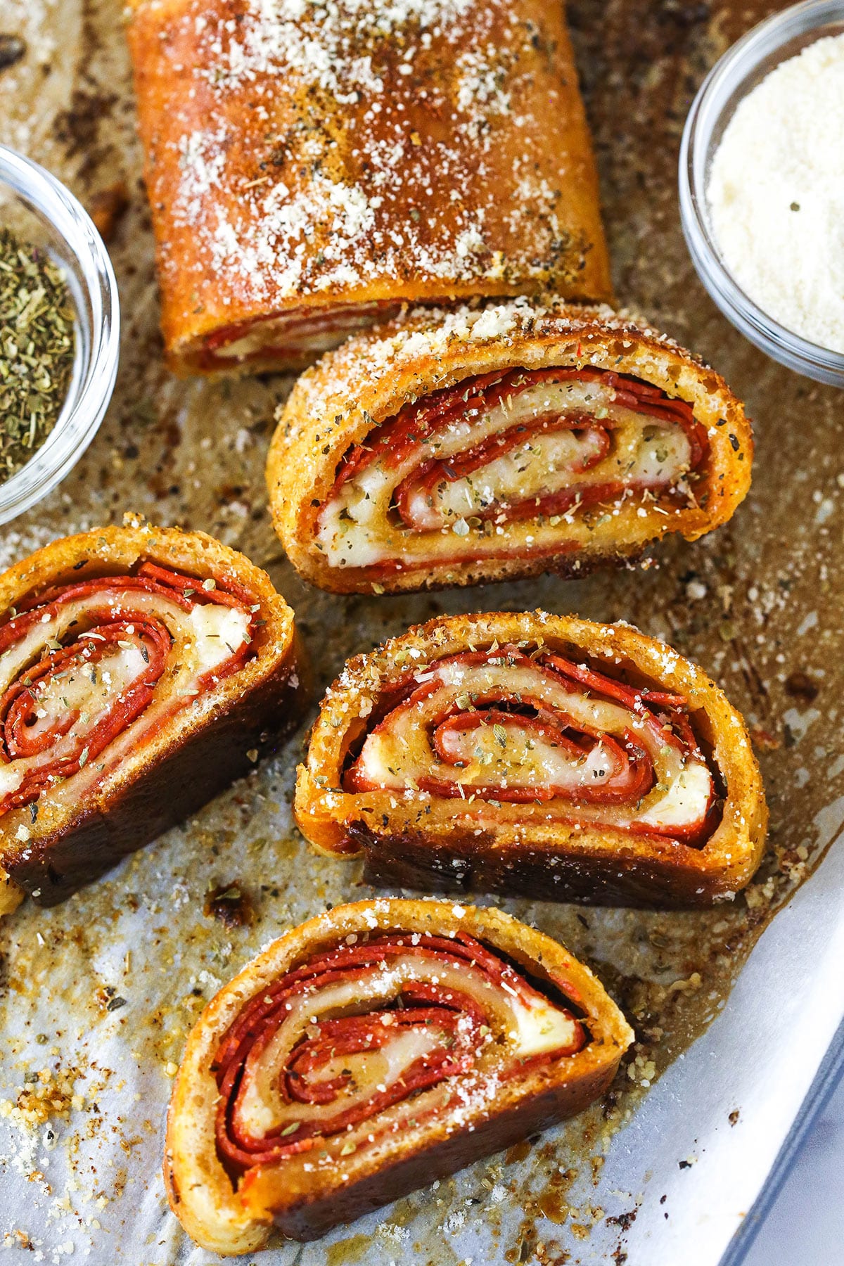 A full pepperoni roll with slices of pepperoni roll near a small bowl of Italian seasoning and a small bowl of cheese.