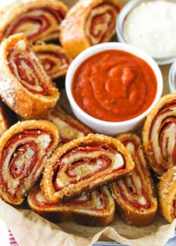 Closeup of pepperoni rolls with marinara sauce and parmesan cheese on a serving tray lined with parchment paper.