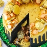 Overhead view of a Pot Of Gold Cake with a slice taken out and decorated with gold wrapped chocolate coins and gold drip in a black pan