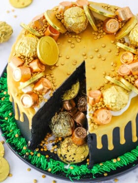 Overhead view of a Pot Of Gold Cake with a slice taken out and decorated with gold wrapped chocolate coins and gold drip in a black pan