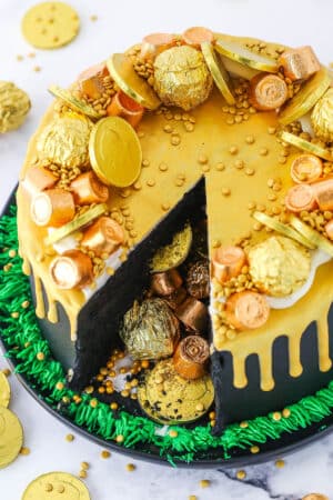 Overhead view of a Pot Of Gold Cake with a slice taken out and decorated with gold wrapped chocolate coins and gold drip in a black pan