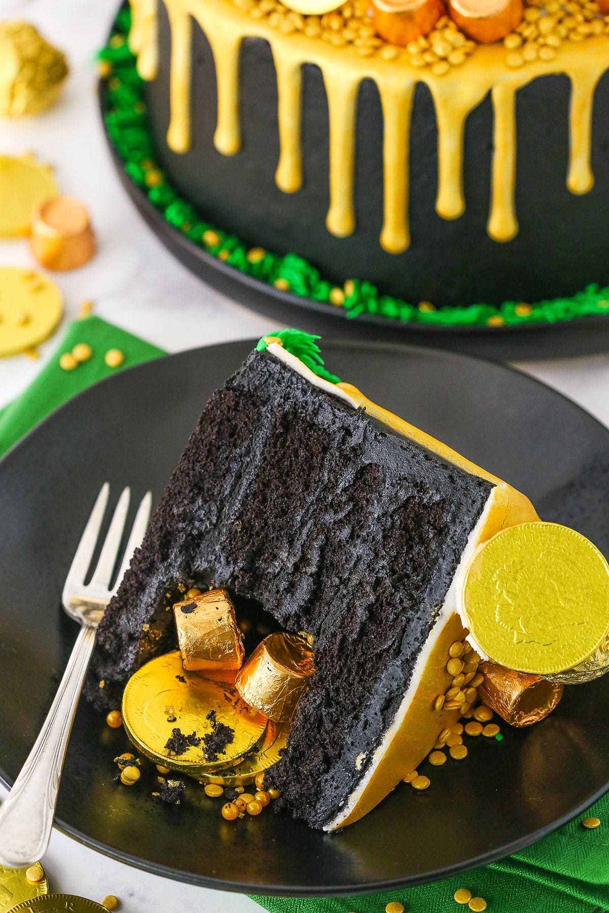 A slice of Pot Of Gold Cake decorated with gold wrapped chocolate coins and gold drip on a black plate with a fork