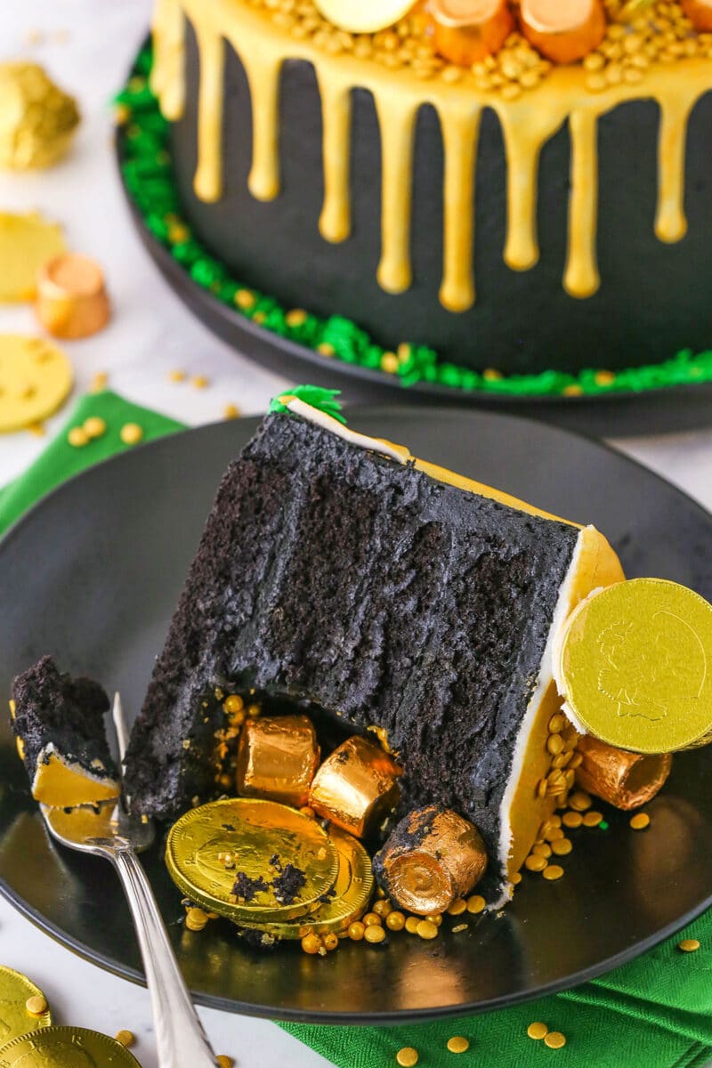 A slice of Pot Of Gold Cake decorated with gold wrapped chocolate coins and gold drip with a bite taken out on a black plate with a fork
