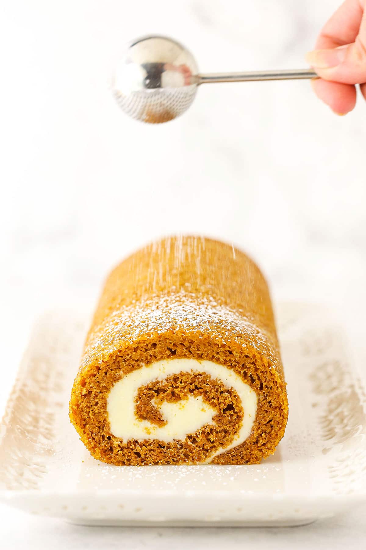 Side view of a Pumpkin Roll Cake in a white platter and powdered sugar being sprinkled on top.