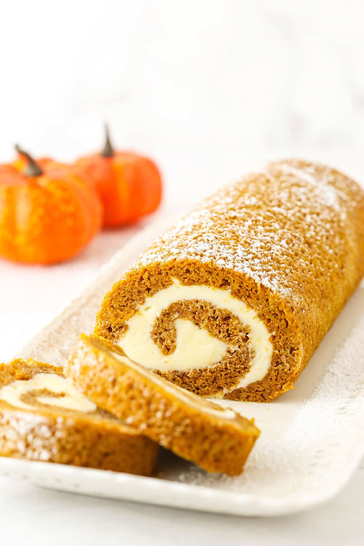 This pumpkin roll recipe is made with tender pumpkin cake, a heavenly cream cheese filling, and easy-to-follow, no-fuss directions. Two slices of Pumpkin Roll Cake layered in front of the rest of the Pumpkin Roll Cake on a white platter.