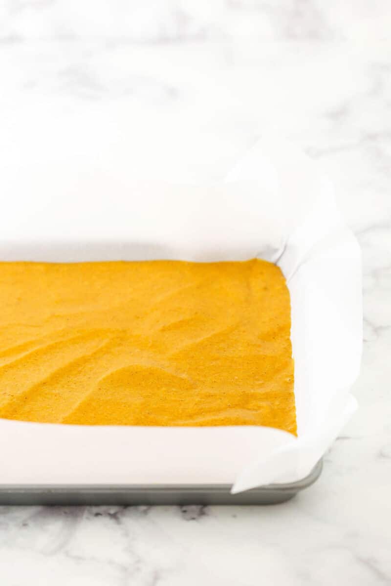 A step in making Pumpkin Roll Cake showing the cake batter being poured into a cake pan lined with parchment paper.