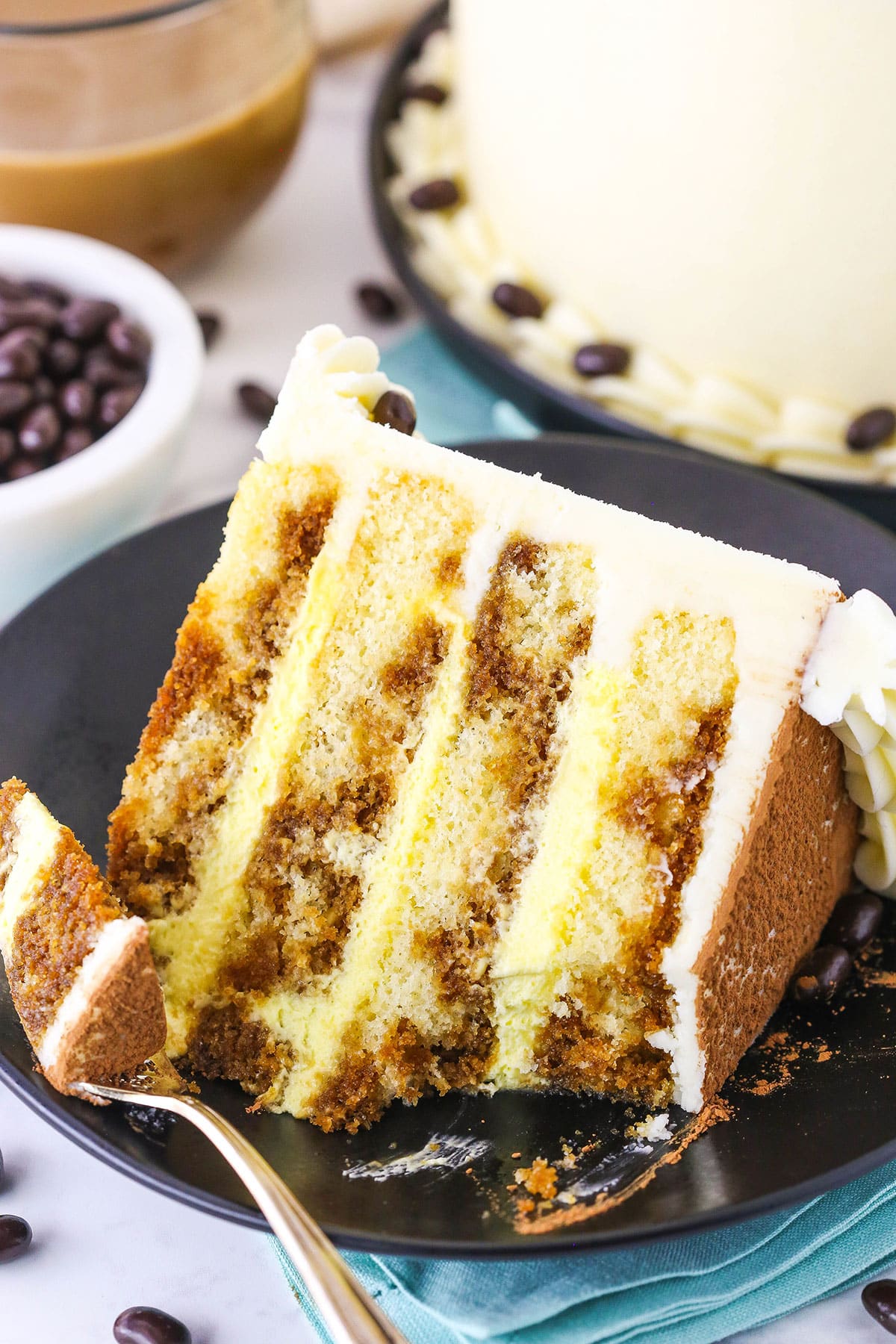 A slice of Tiramisu Layer Cake with a bite removed on a black plate with a fork