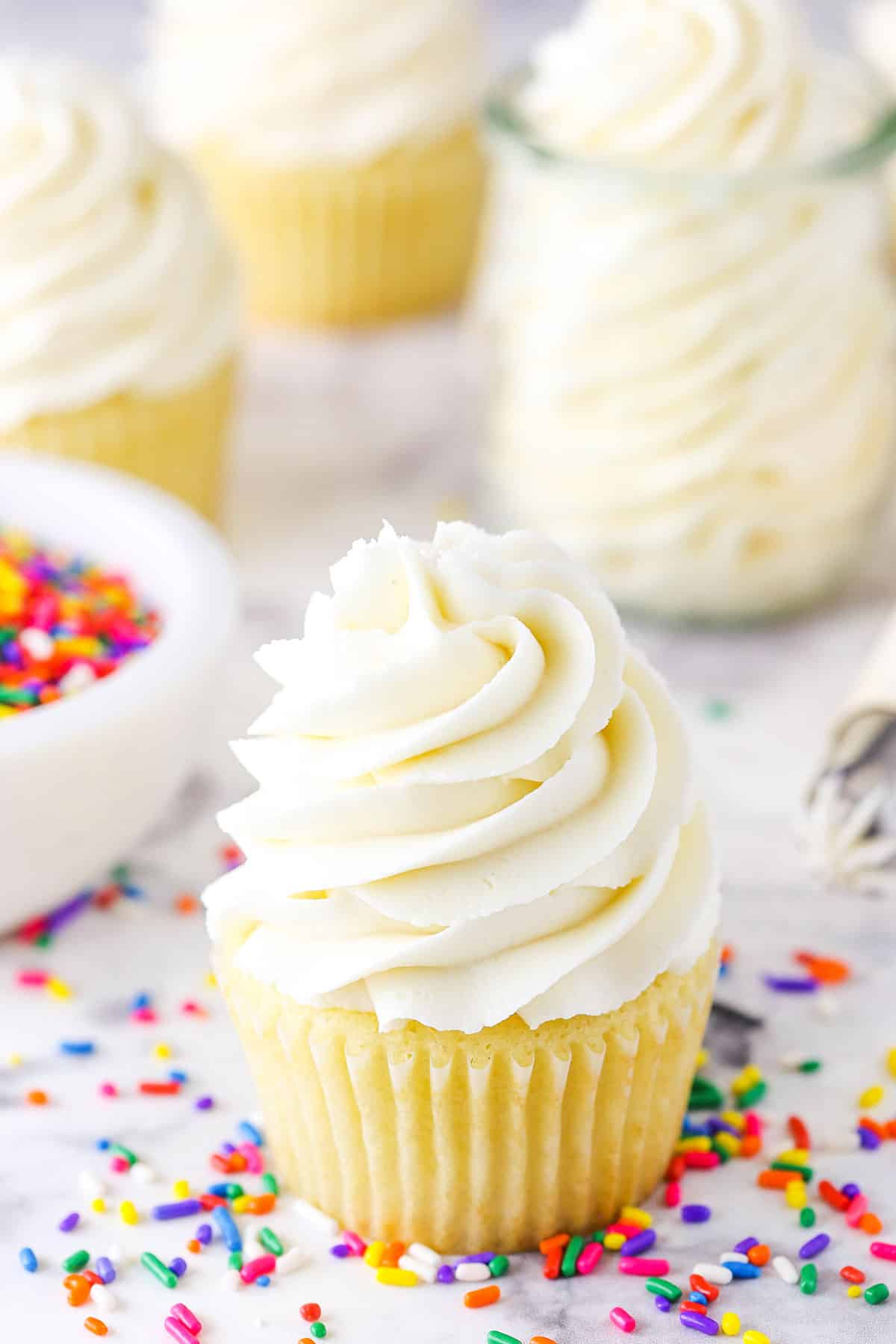 Vanilla Buttercream Frosting piped onto a vanilla cupcake surrounded by multicolored sprinkles