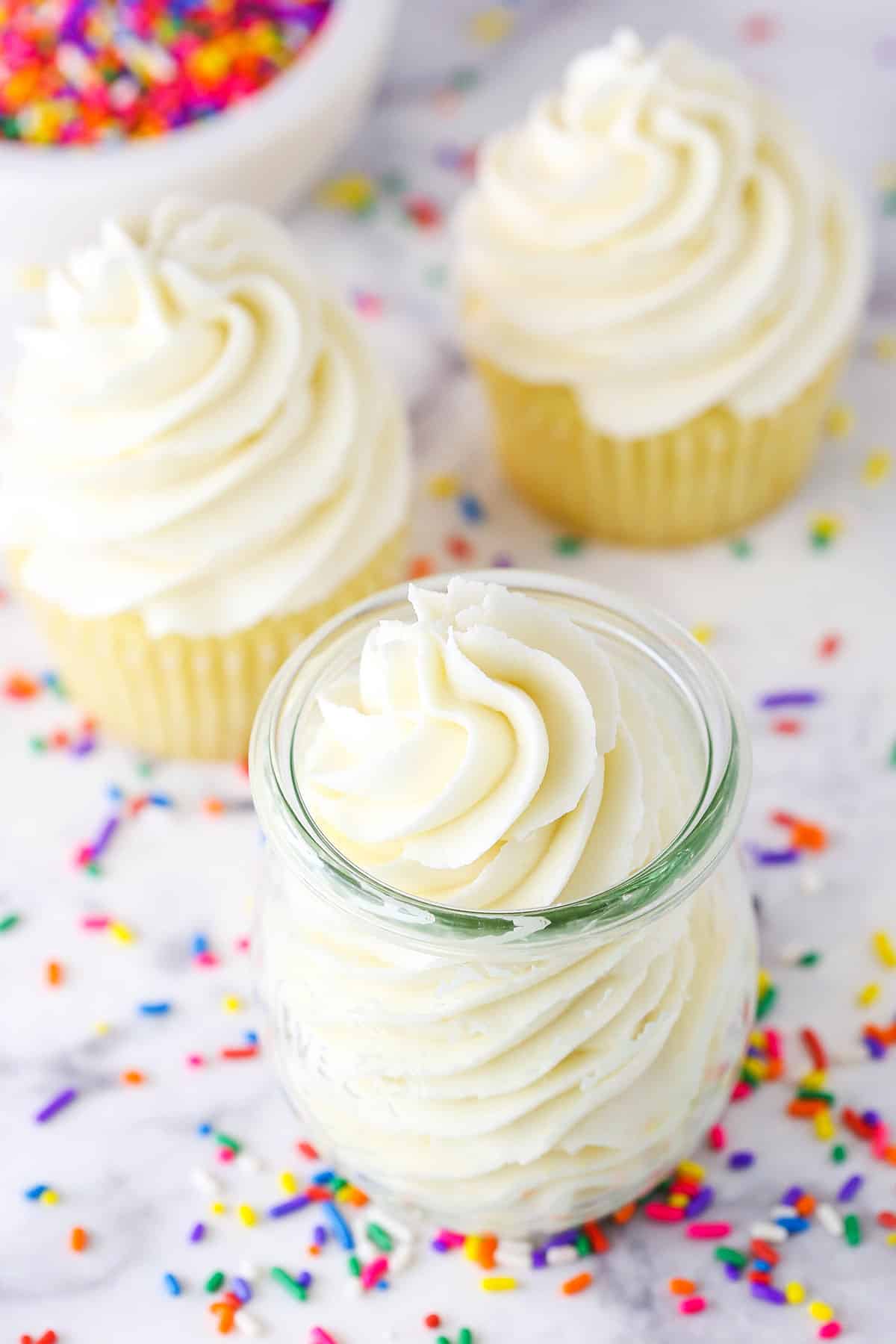 Vanilla Buttercream Frosting piped into a glass jar on a white marble table top surrounded by multicolored sprinkles