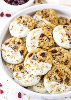 White Chocolate Dipped Cranberry Oatmeal Cookies coated with chopped pecans layered in a white serving platter with a small dish of dried cranberries in the background