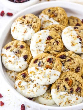 White Chocolate Dipped Cranberry Oatmeal Cookies coated with chopped pecans layered in a white serving platter with a small dish of dried cranberries in the background