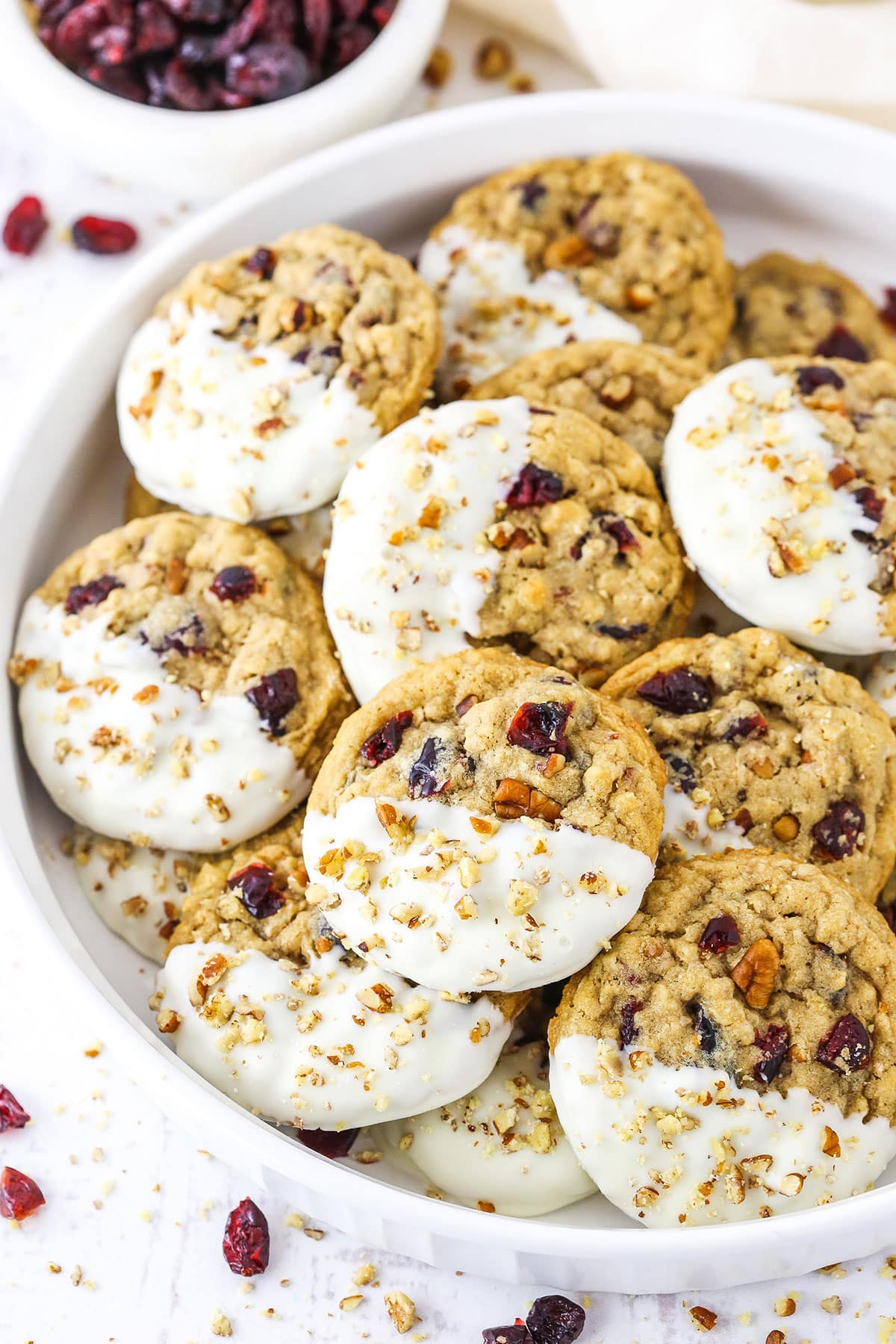 White Chocolate Dipped Cranberry Oatmeal Cookies coated with chopped pecans layered in a white serving platter with a small dish of dried cranberries in the background