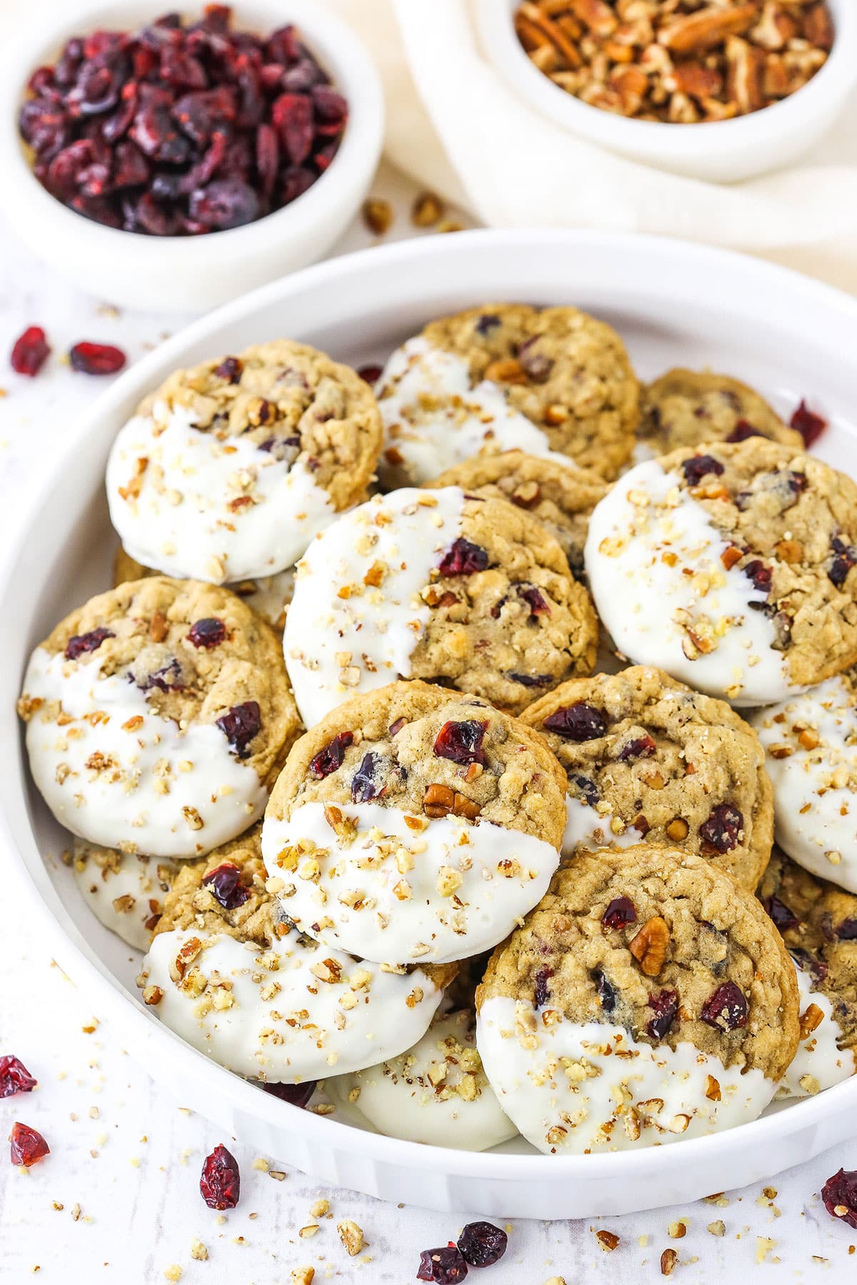 White Chocolate Dipped Cranberry Oatmeal Cookies coated with chopped pecans layered in a white serving platter with dried cranberries and chopped pecans in the background