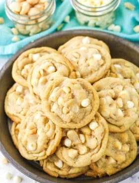 White Chocolate Macadamia Nut Cookies stacked in a dark platter with white chocolate chips and macadamia nuts in the background