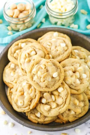 White Chocolate Macadamia Nut Cookies stacked in a dark platter with white chocolate chips and macadamia nuts in the background